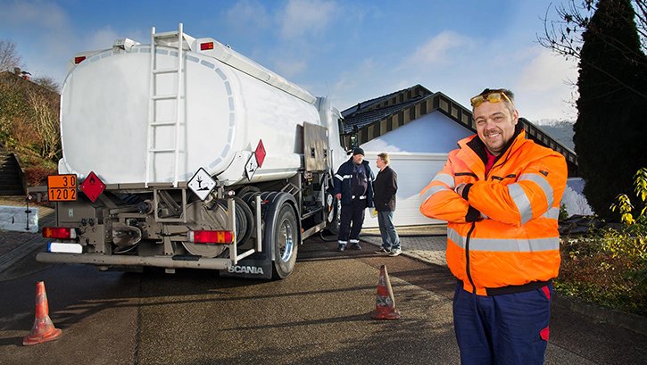 Im Vordergrund ein Mann mit Brille. Im Hintergrund ein Heizöl-Tankwagen mit zwei Personen und eine Garage
