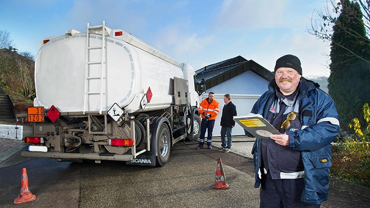 Im Vordergrund ein Mann mit einem Klemmbrett. Im Hintergrund ein Heizöl-Tankwagen mit zwei Personen und eine Garage